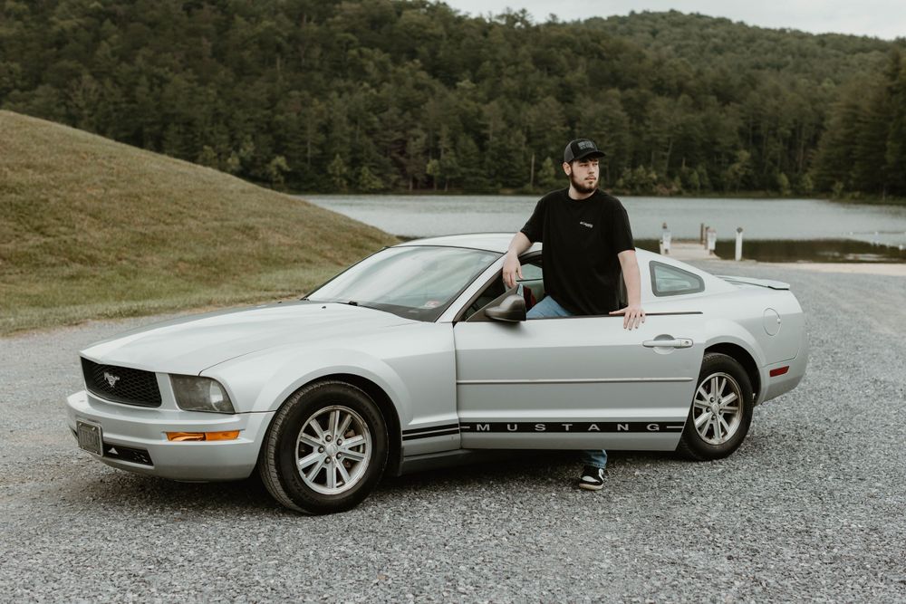 A silver Mustang sports car parked near a lake with hills in the background.