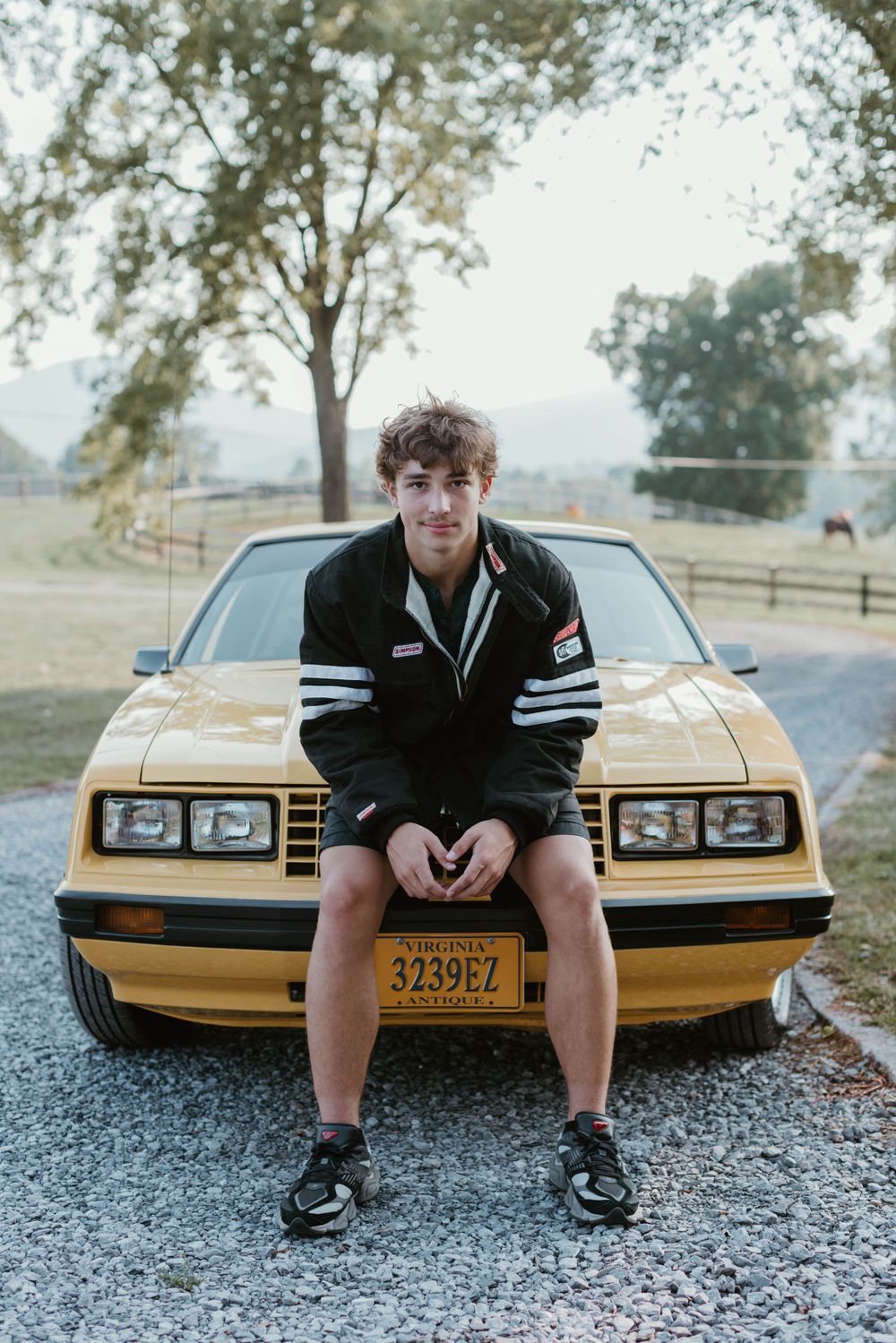 A person in athletic wear sits on hood of gold vintage Mustang on gravel road.
