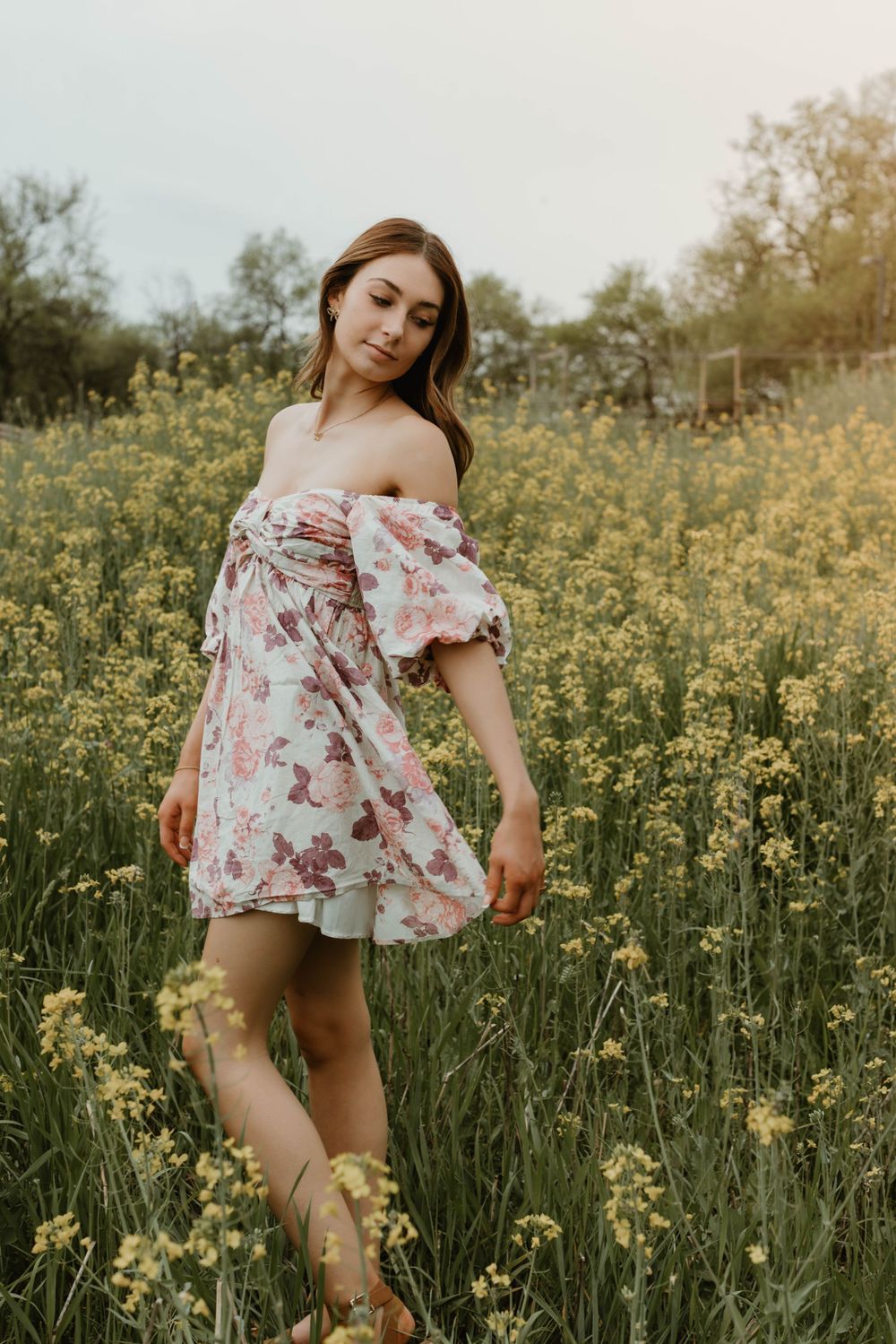 Woman in floral dress poses playfully in field of yellow wildflowers during golden hour.
