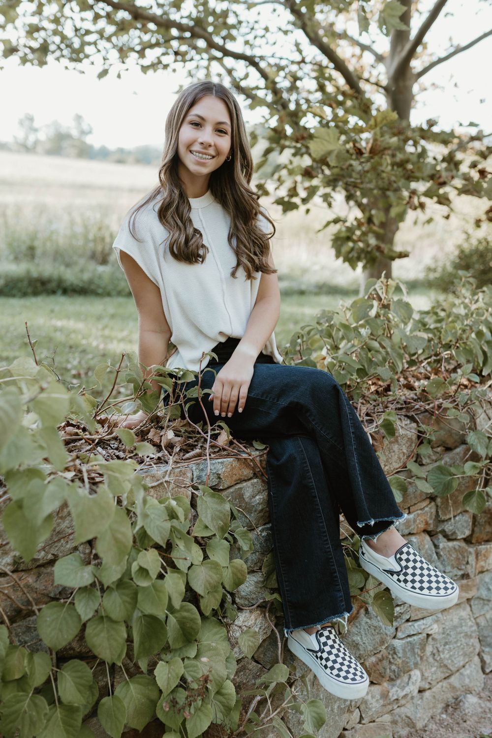 A casual portrait of a woman sitting on stone wall wearing checkered vans and wide-leg jeans.