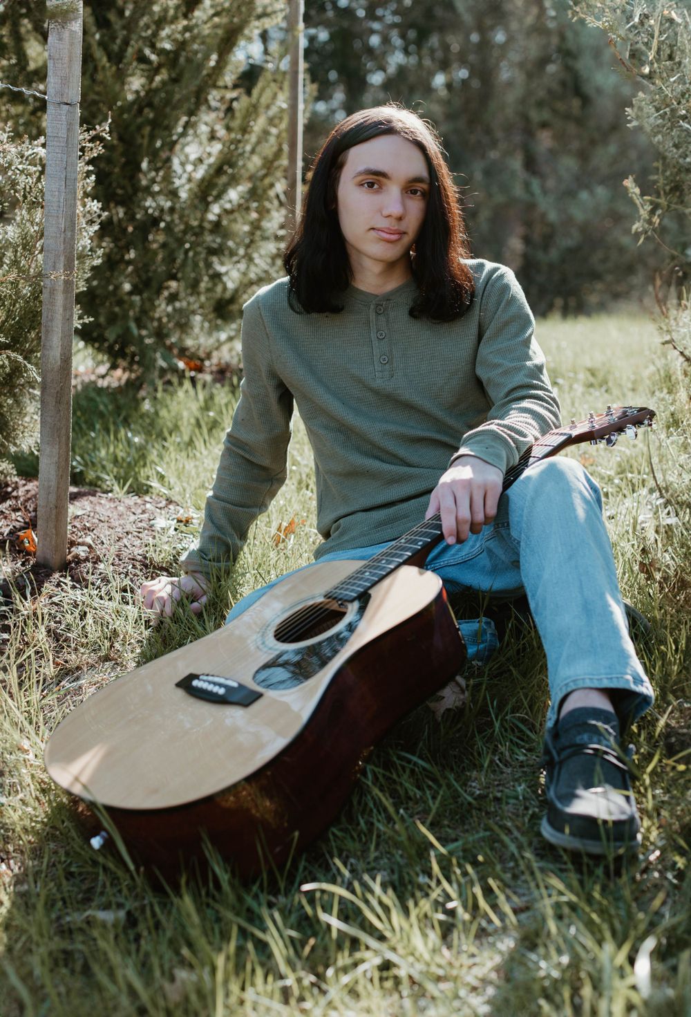 Person in casual wear sits in grass with acoustic guitar during outdoor photo session.