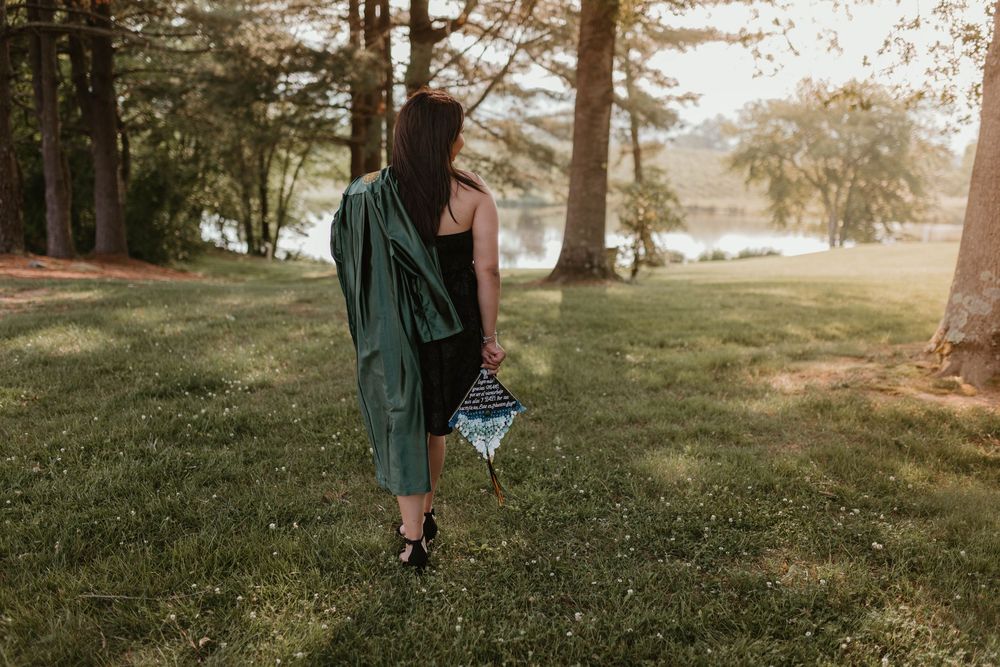 Graduate in green cap and gown walks through a sunlit park during golden hour.
