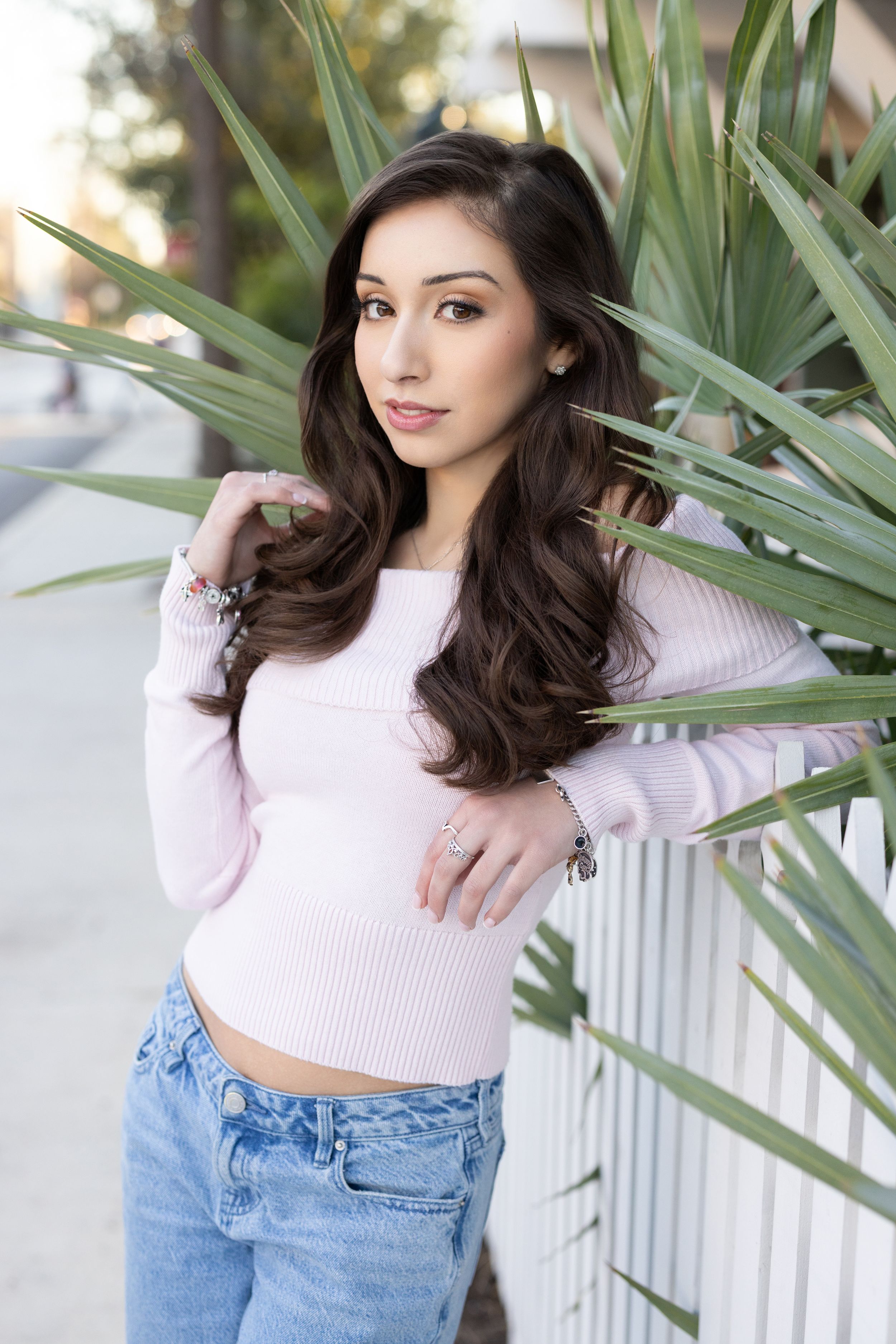 A person in a pink crop top sweater and jeans poses against a white fence with tropical plants in the background.