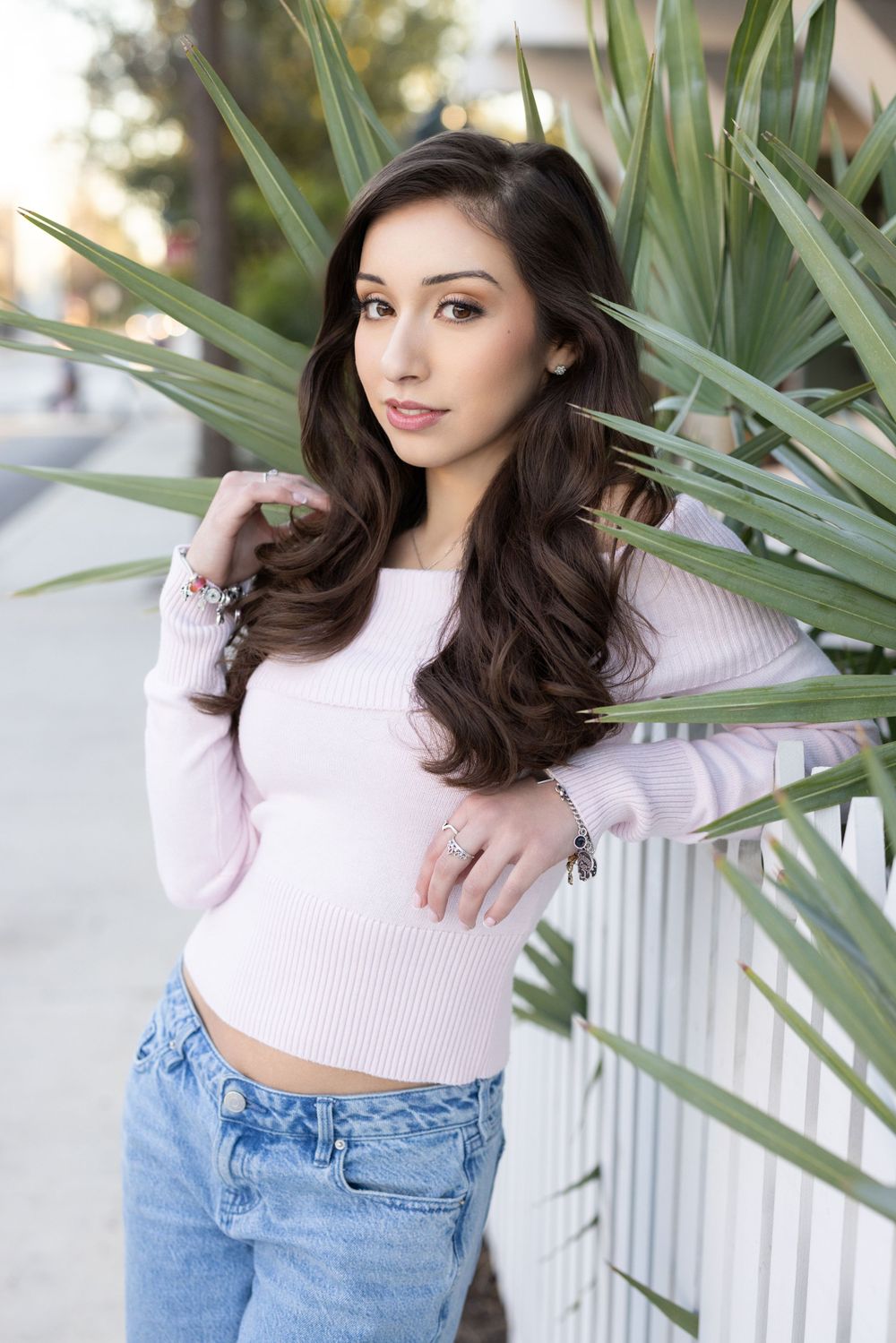 A person in a pink crop top sweater and jeans poses against a white fence with tropical plants in the background.