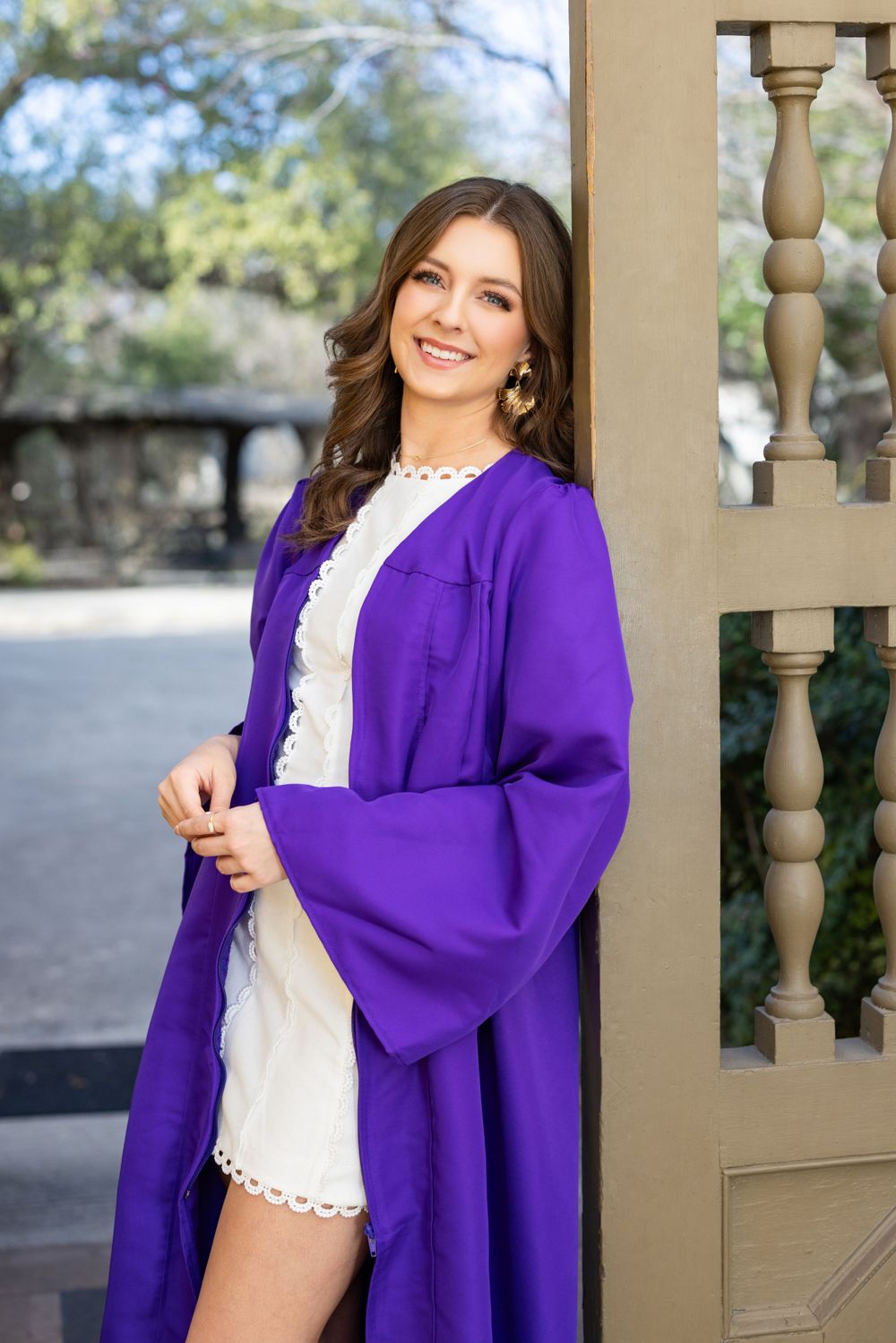 Graduate wearing a bright purple graduation gown and white lace dress poses smiling by a beige railing outdoors.