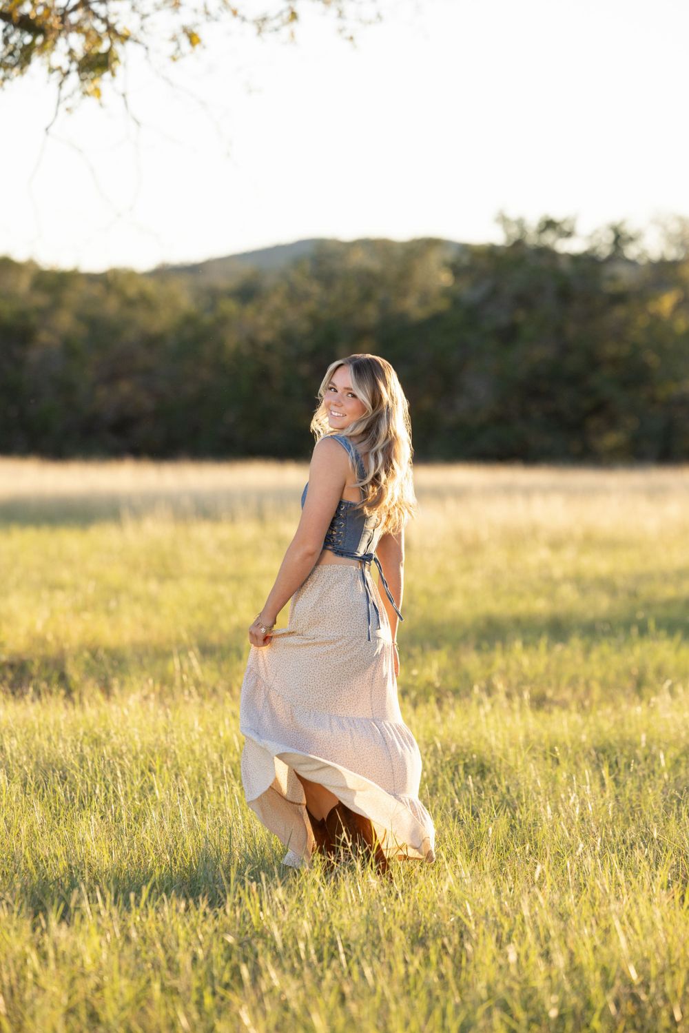 A woman in a flowing white skirt twirls in a golden sunlit meadow during sunset with mountains in the background.