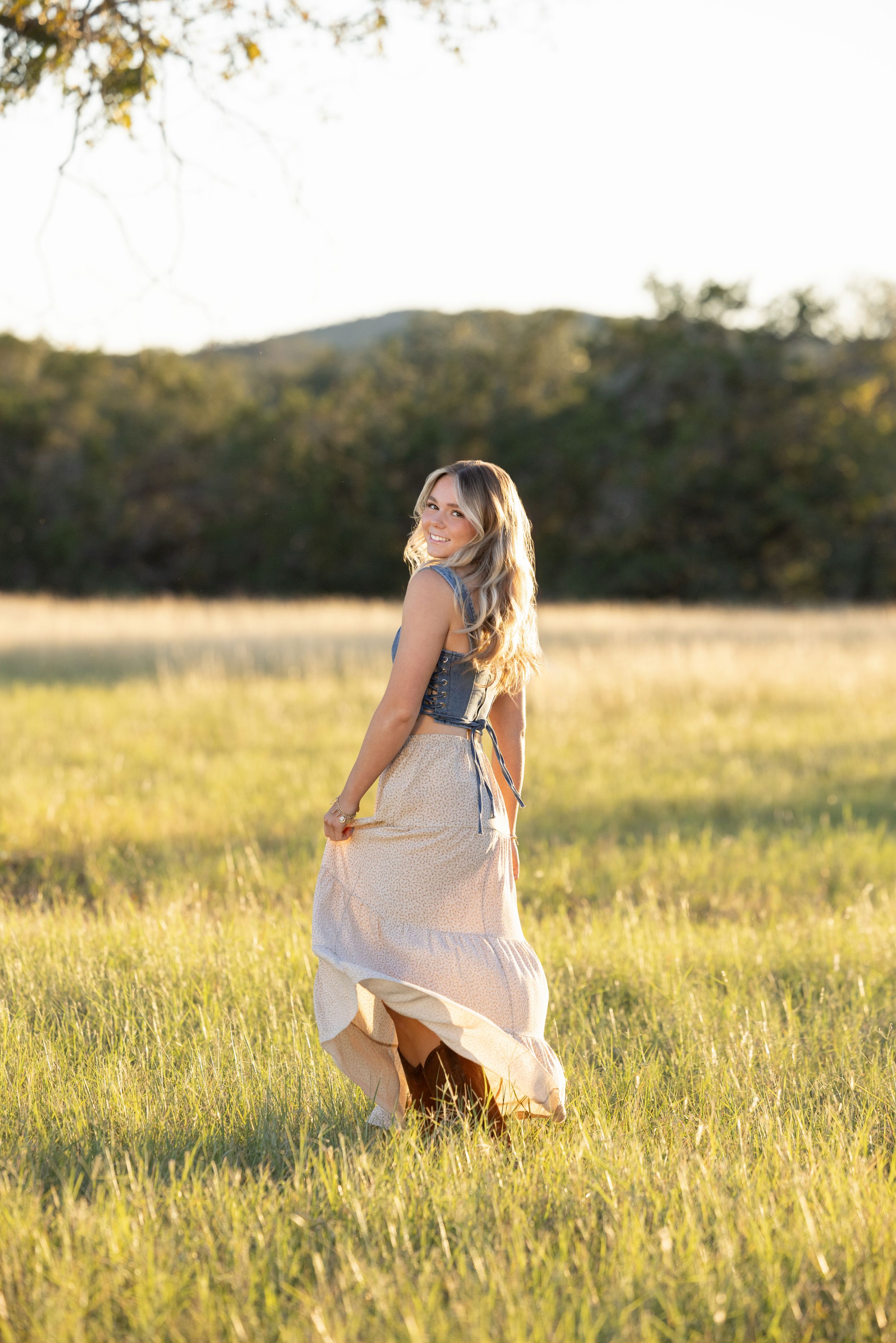 A woman in a flowing white skirt twirls in a golden sunlit meadow during sunset with mountains in the background.