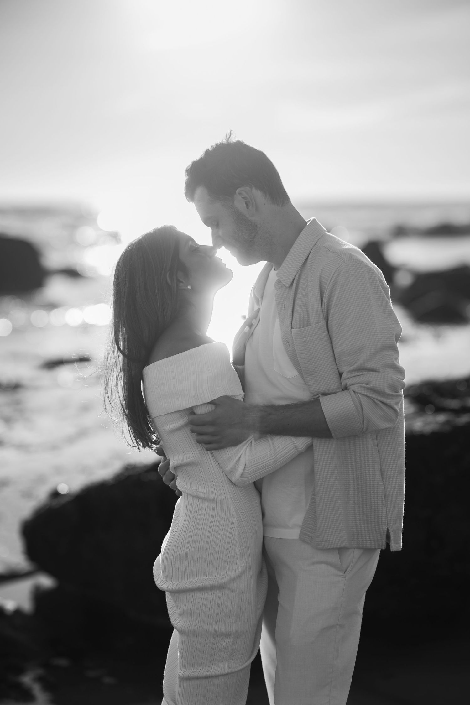 Romantic couple embracing and kissing at sunset on a rocky beach in black and white.
