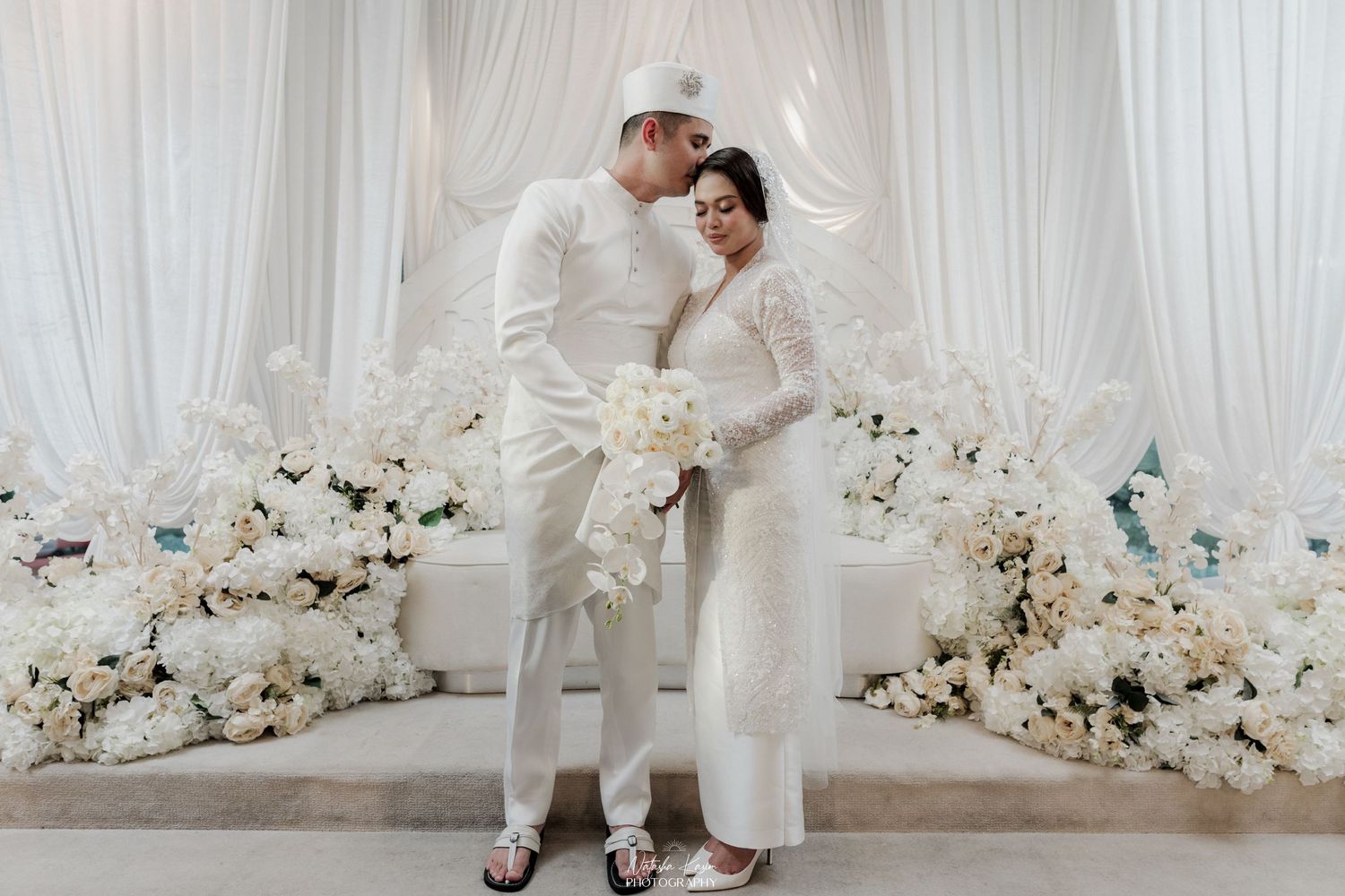 A couple in white outfits poses against an elegant white backdrop with floral arrangements.