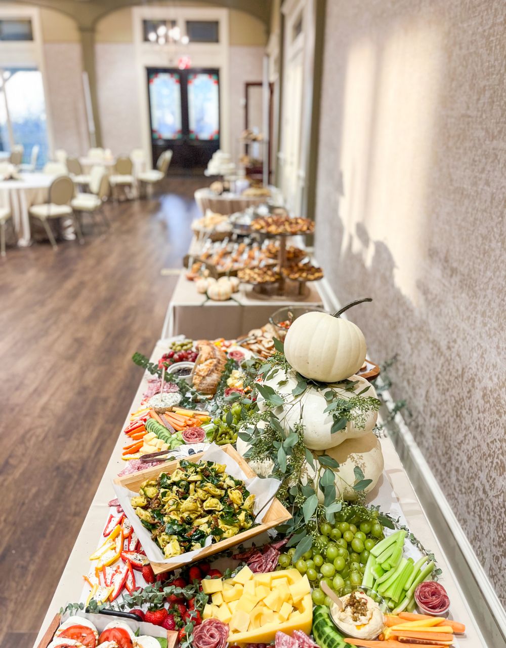Long grazing table with white pumpkins, fresh vegetables, and assorted appetizers in a bright venue.