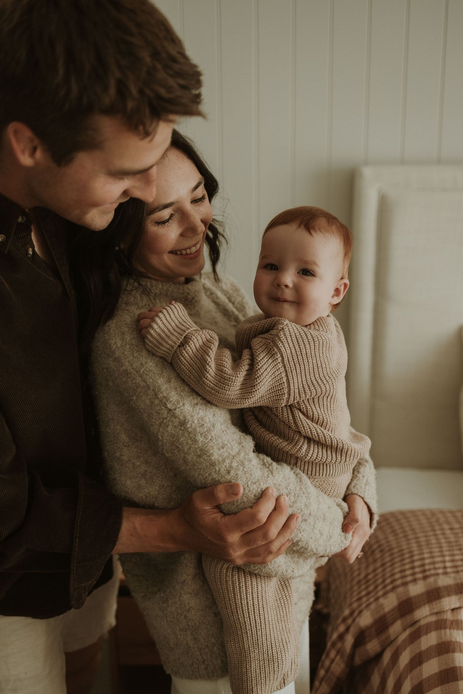 A young family shares a tender moment, parents smiling at their happy baby in a cozy, warmly lit room.