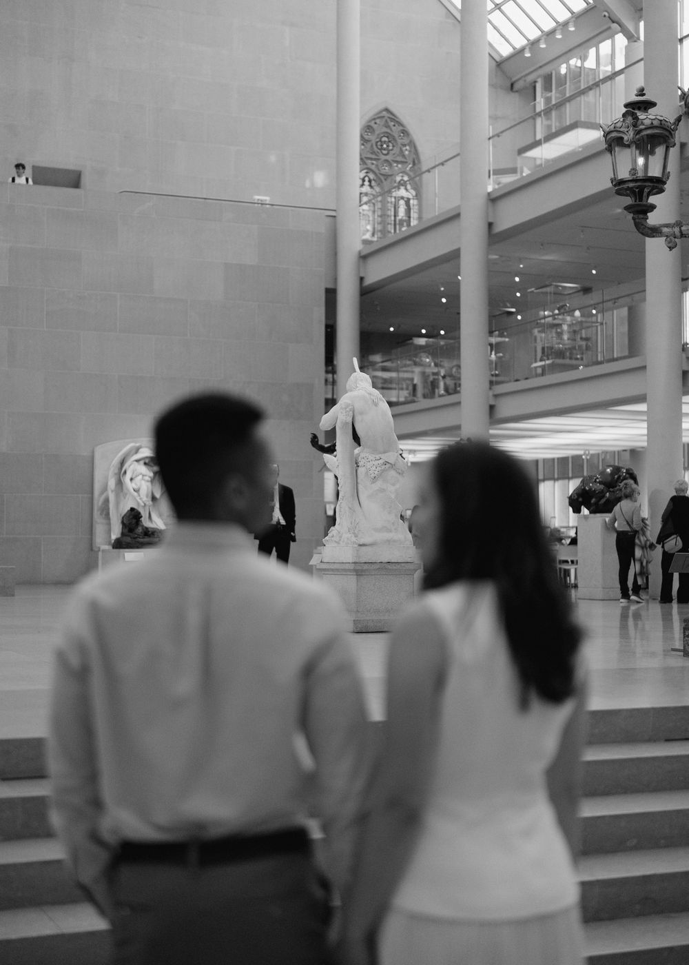 Couple views classical sculpture exhibit in grand museum atrium, black and white photography.