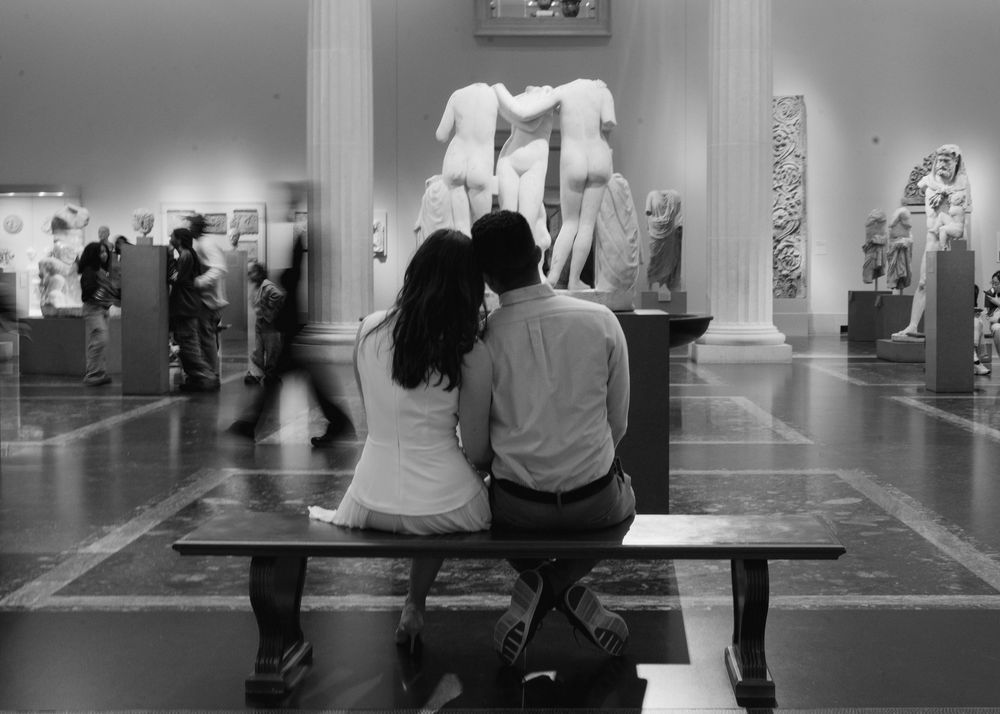 Couple sits intimately on bench admiring classical sculptures in busy art museum, black and white.