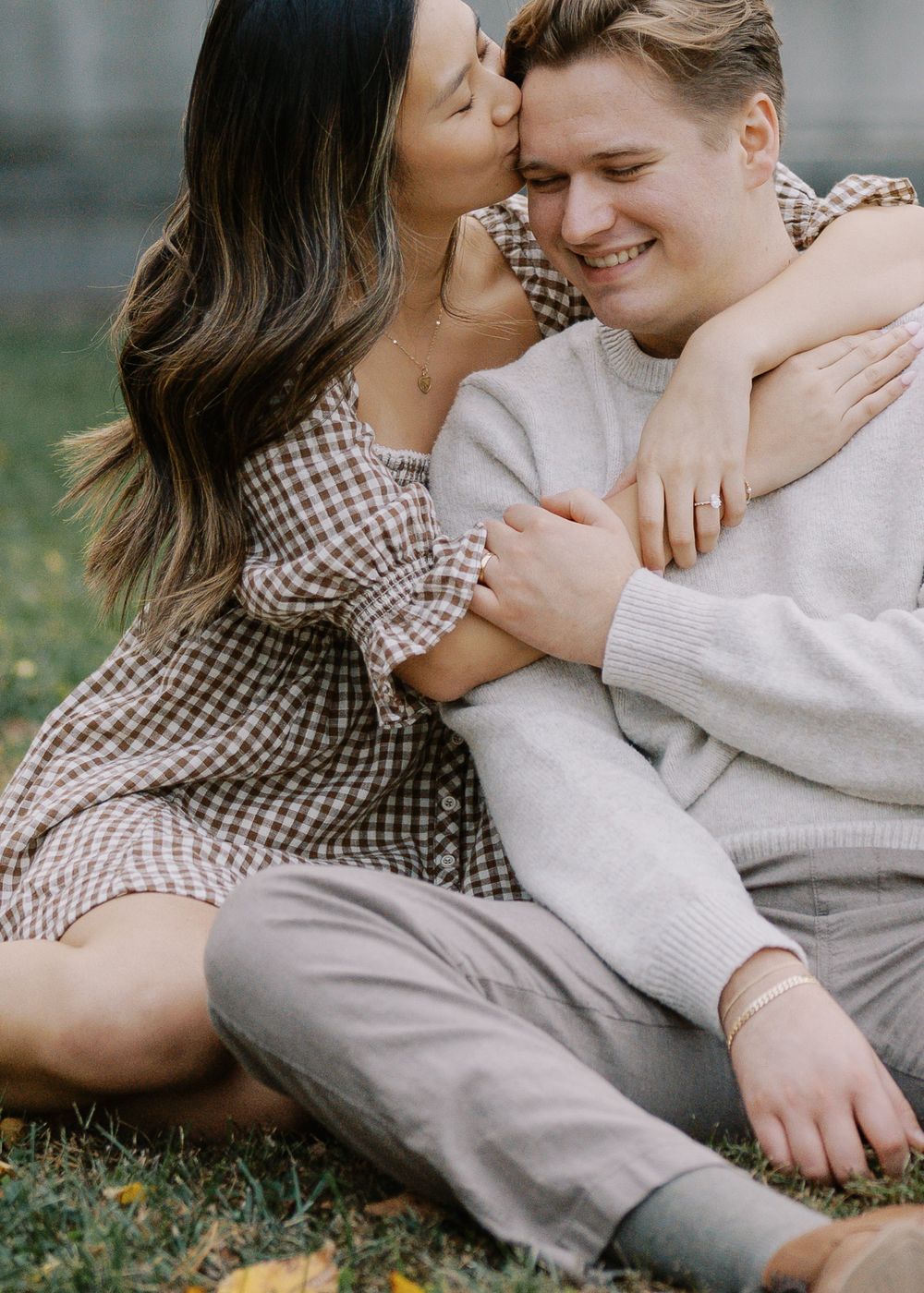 Woman kissing her smiling fiance on the cheek while sitting together on grass outdoors.