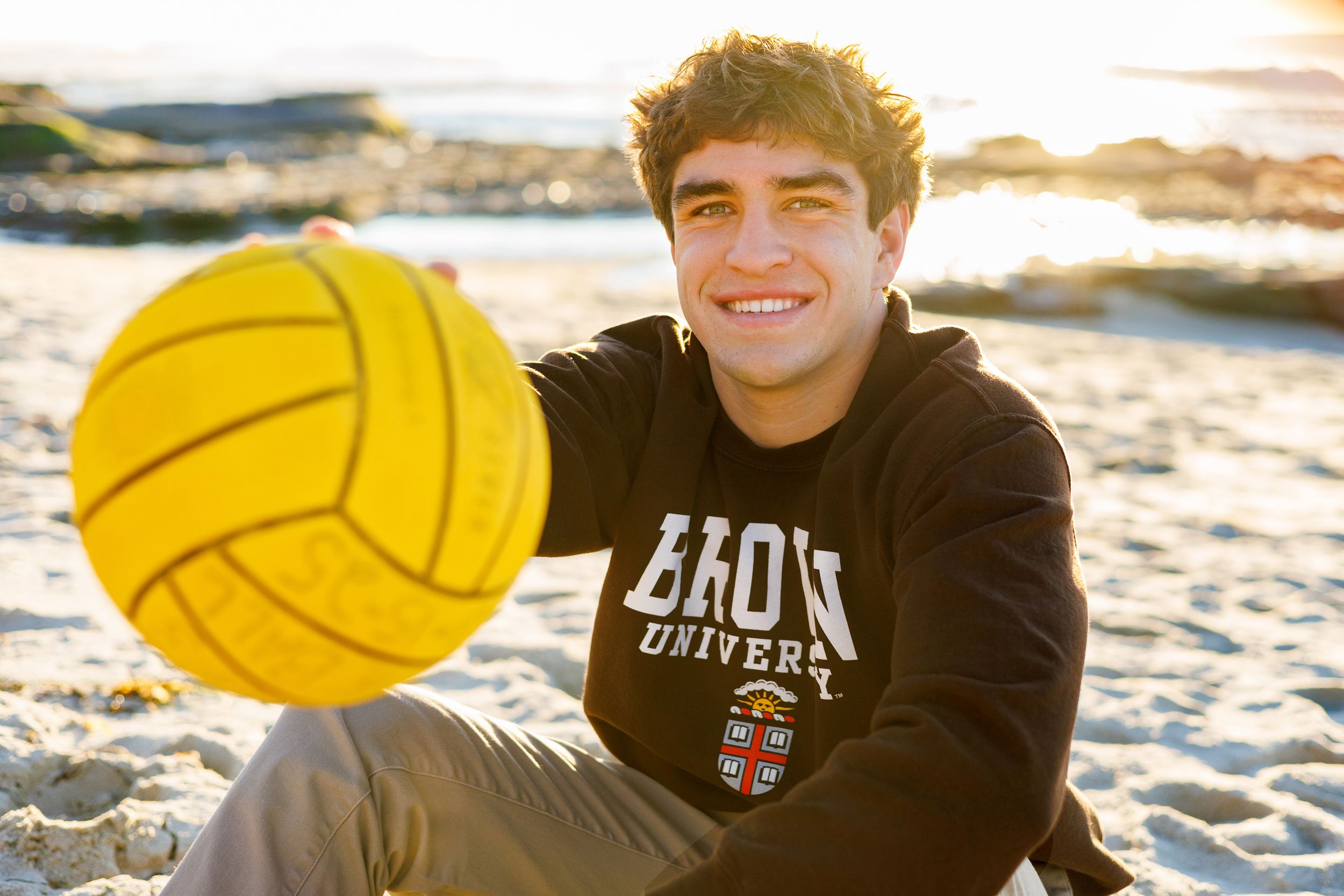 Young man in Brown University hoodie smiling on beach with yellow water polo ball.