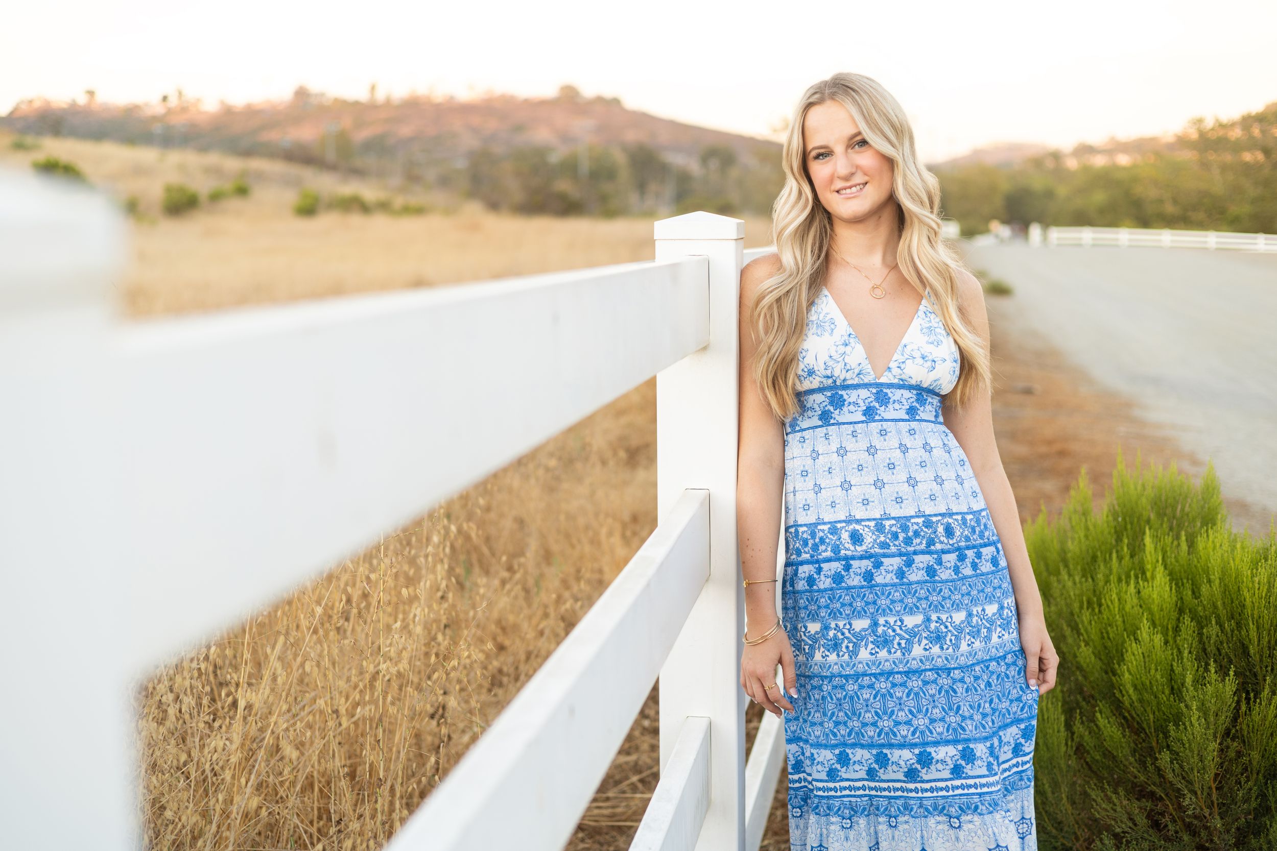 Woman in blue floral maxi dress leaning on white fence in sunny outdoor setting.