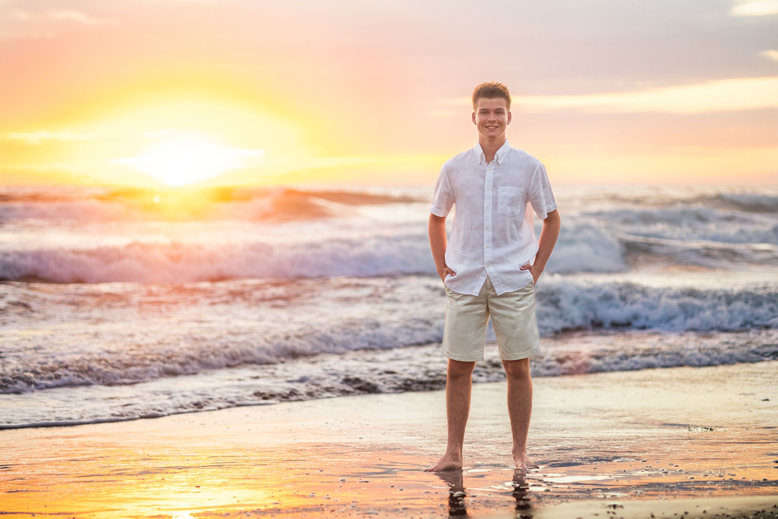 Young man in a white shirt and khaki shorts stands barefoot on a wet sandy beach as waves roll in under a glowing orange sunset.
