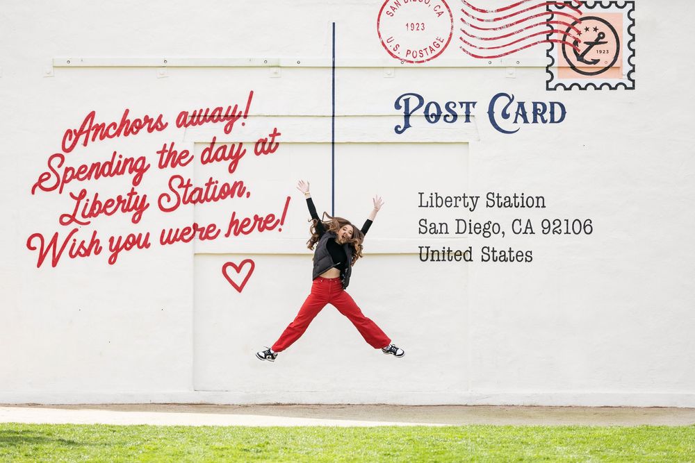 A person in red pants jumps joyfully against a white wall with a postcard-style message about Liberty Station in San Diego.