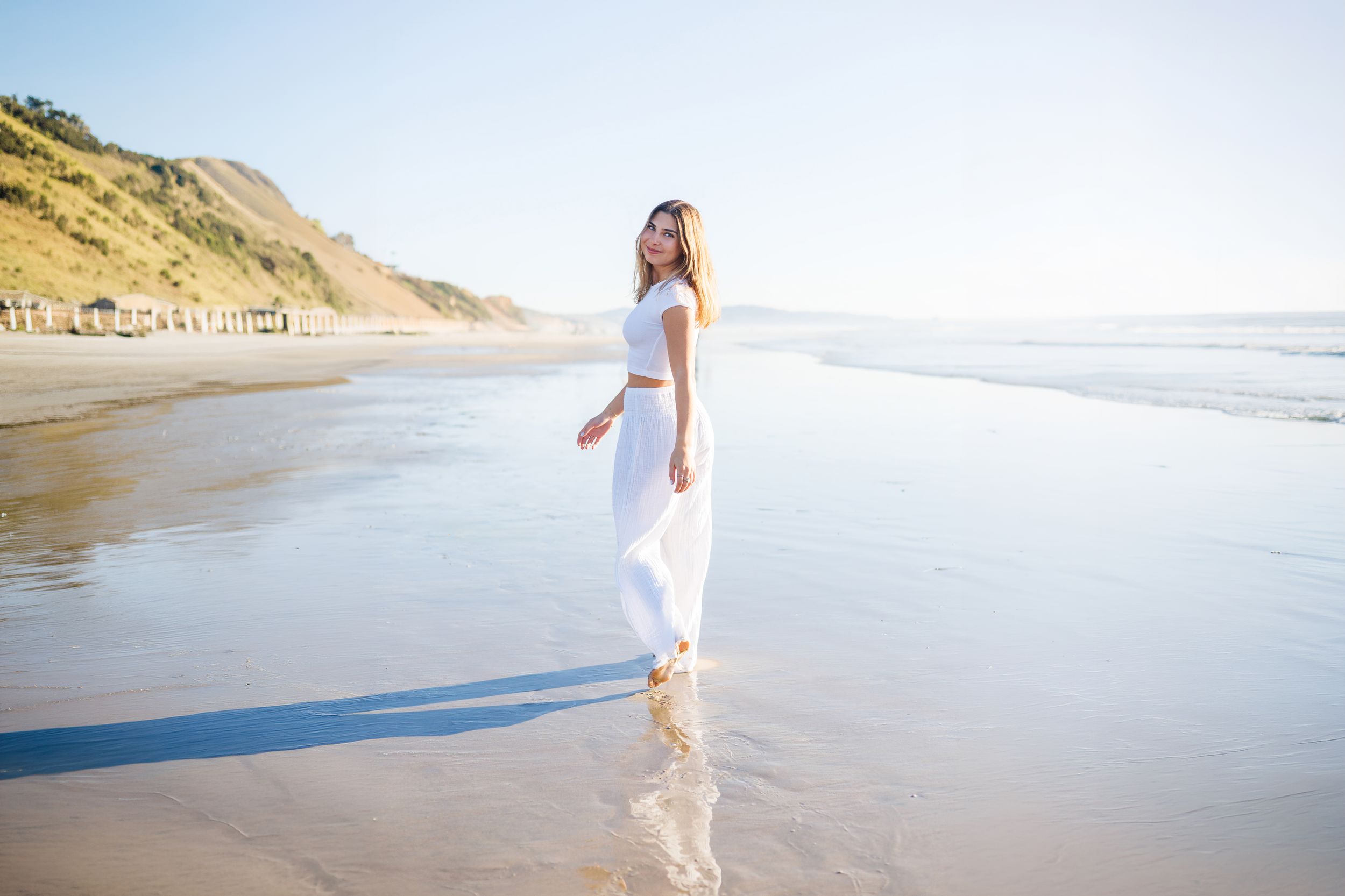 Woman in white outfit walking on sunny beach with ocean waves and cliffs.