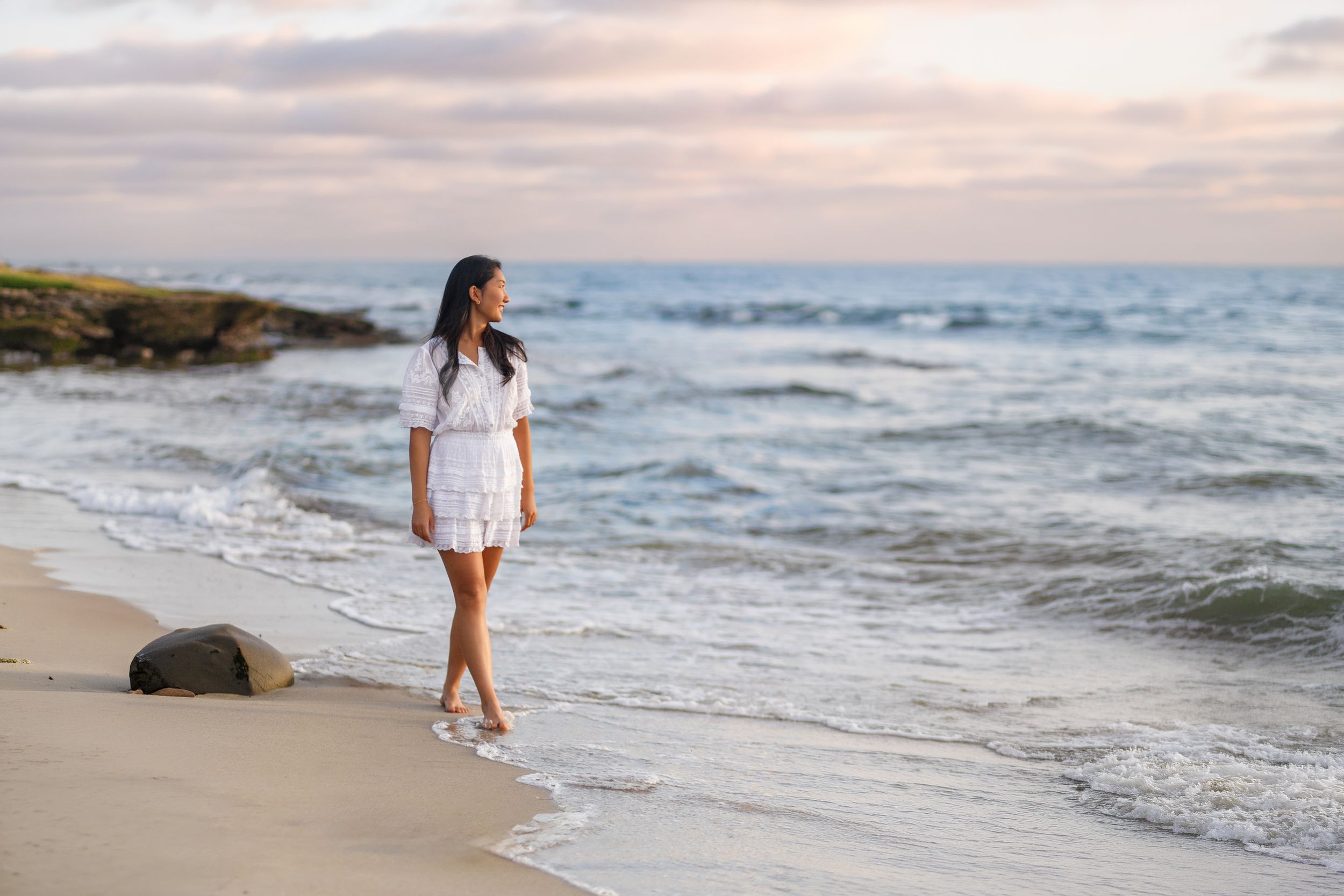 Woman in white dress walking barefoot on a sandy beach at sunset with ocean waves in the background.