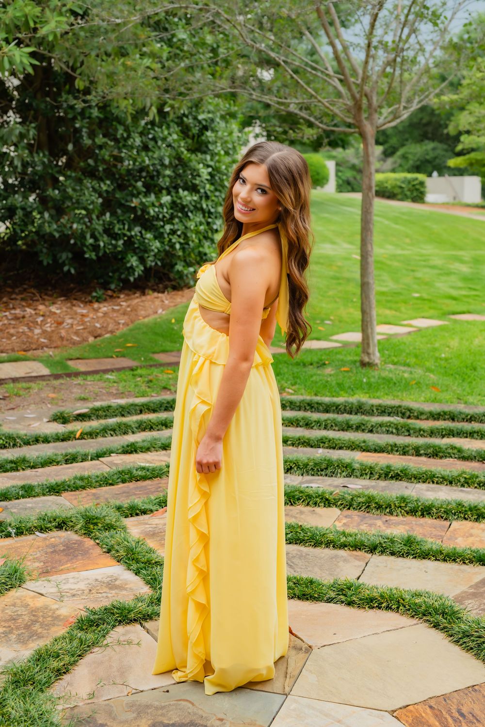 Woman in yellow maxi dress posing on stone path in lush garden setting.