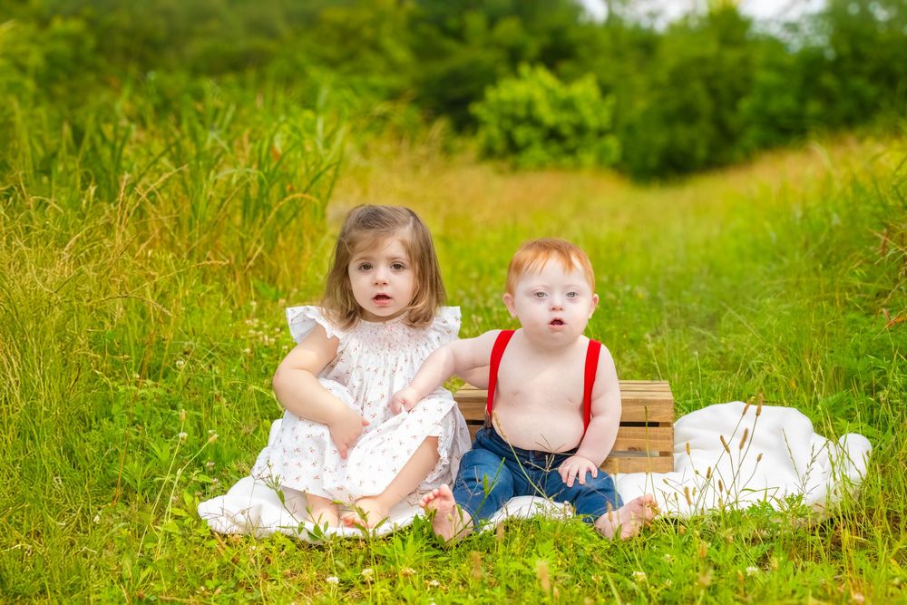 Two young children sit together on a white blanket in a sunny meadow with tall grass and greenery.
