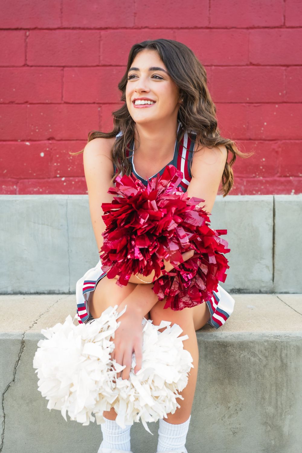 Cheerleader with red and white pom poms smiles while sitting against red brick wall.