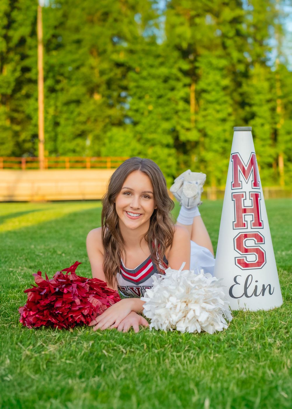 A cheerleader in red and white uniform poses with pom poms and a customized megaphone on a grassy sports field.