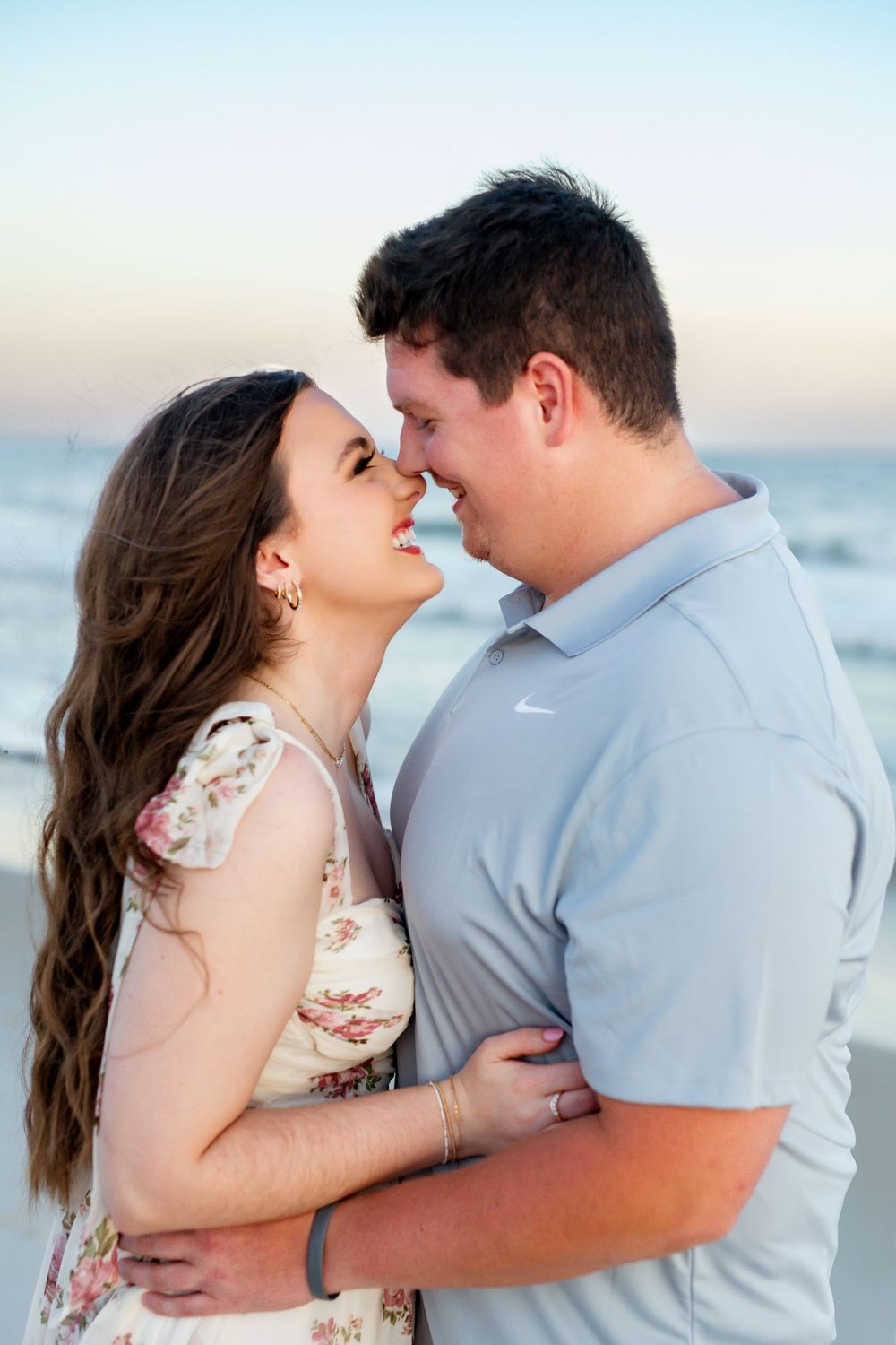 A romantic couple in casual attire embraces while touching noses at the beach during sunset.