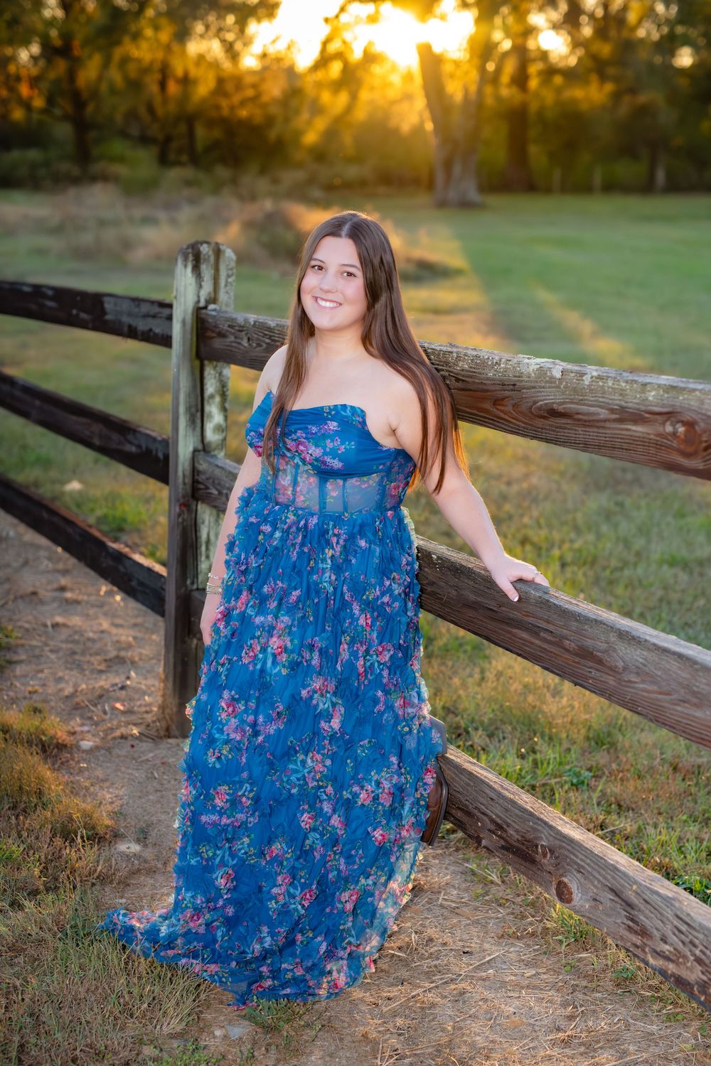 A person in a flowing blue floral dress poses gracefully against a rustic wooden fence at sunset.