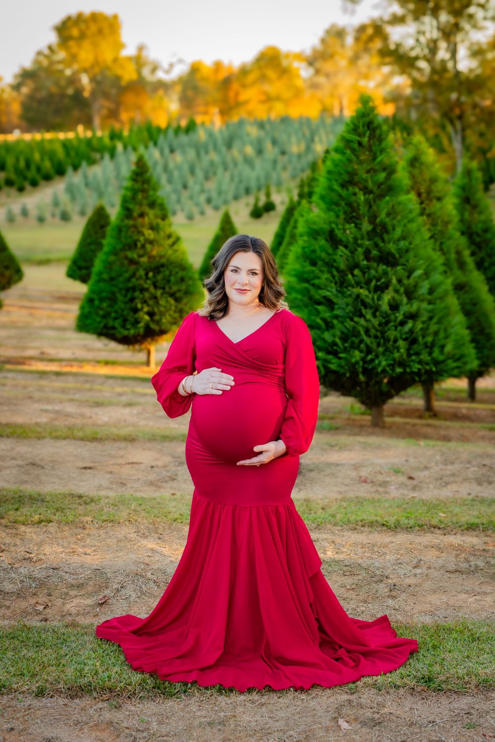 Expectant woman in flowing red gown poses elegantly at Christmas tree farm with autumn colors in background.