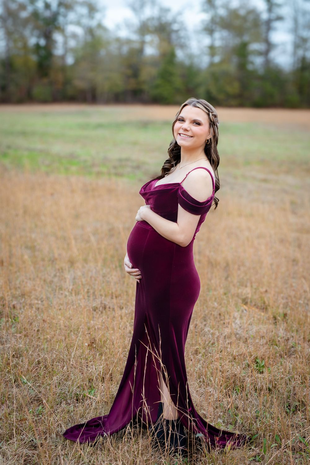 A pregnant woman in a burgundy velvet dress poses gracefully in a golden autumn field at sunset.