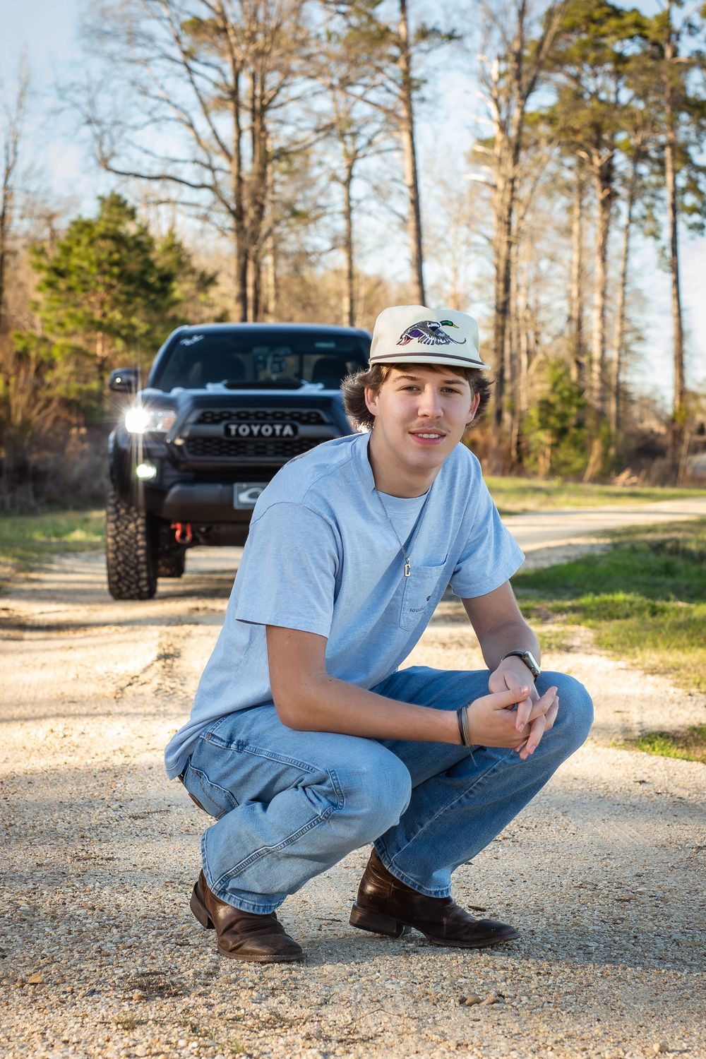 Young man in cap and jeans crouching on dirt road with Toyota Tacoma truck in background.