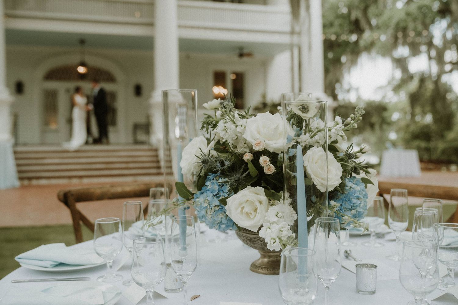 Elegant wedding reception table setting with white and baby blue floral centerpiece and crystal glassware on outdoor patio.