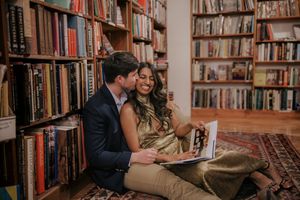Couple sitting close together browsing books in cozy library with wooden shelves and warm lighting.