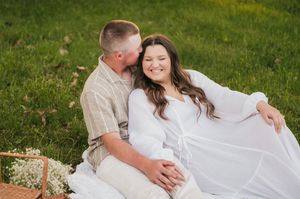 A romantic couple sitting on grass during an outdoor photoshoot, wearing white and neutral colors in a natural setting.