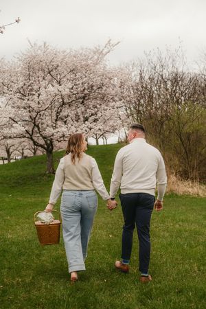 A couple walking through a cherry blossom orchard carrying a picnic basket on a spring day.
