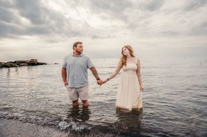 A couple holds hands while standing in shallow ocean water at sunset against a cloudy sky and rocky coastline.