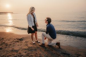 A couple shares a romantic marriage proposal moment on the beach at sunset with ocean waves in the background.