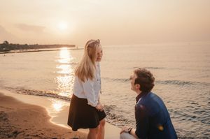 A romantic beach proposal scene at sunset with golden light reflecting on the ocean waves.