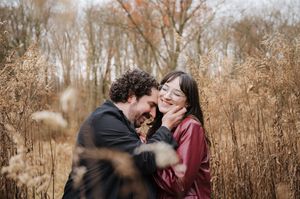 A couple embraces intimately amidst tall brown grass and autumn foliage in a natural outdoor setting.