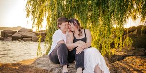 A romantic couple sits together under a weeping willow tree at sunset by the water's edge with rocks in the background.