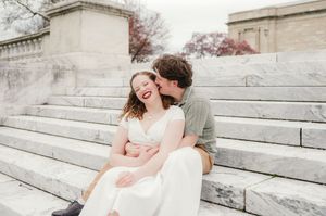 Two people embrace and kiss while sitting on wide stone steps, with one wearing a formal white dress.