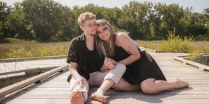 Two people wearing black outfits sit close together on a wooden dock on a sunny summer day.