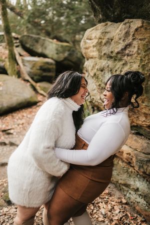 Two people wearing white and brown outfits embrace while sitting together on rocky terrain in a forest setting.