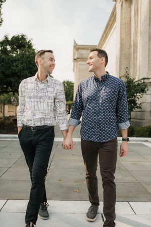 Two people in patterned button-down shirts walking and holding hands on a stone walkway near a building.