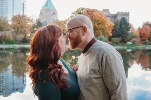 A couple shares a romantic moment by a tranquil lake with autumn trees and a domed building in the background.