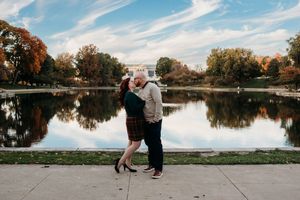 Two people embrace by a serene lake with autumn trees and reflective waters in an urban park setting.