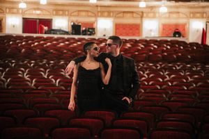 A couple in formal black attire share an intimate moment among empty red velvet theater seats in an elegant auditorium.