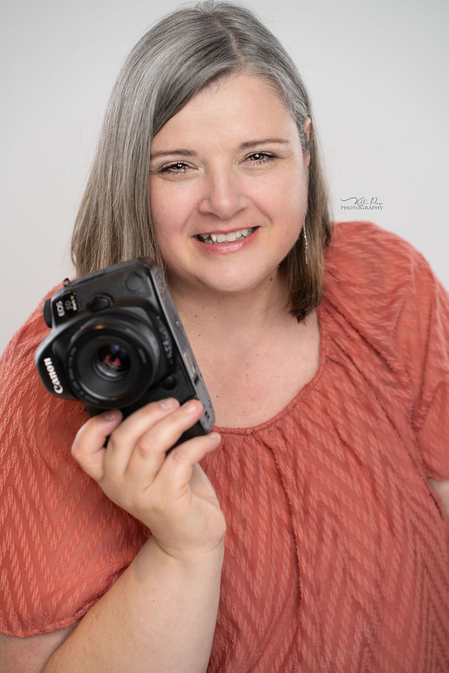 A photographer wearing a coral-colored sweater holds a professional DSLR camera while posing against a light background.